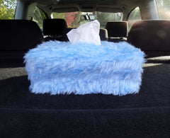 A handmade, rectangular tissue box cover made from soft, fuzzy baby blue faux fur, shown sitting on a dark surface in the back of a car with a white tissue peeking out of the top.